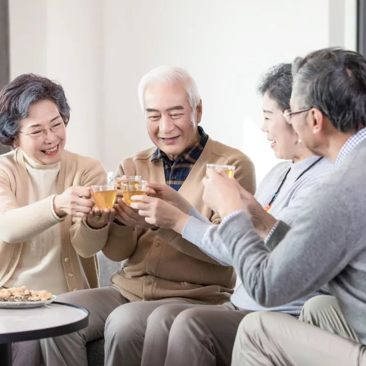 四位长者围坐在一起，手持茶杯，面带微笑，正在进行碰杯庆祝。他们坐在沙发上，面前的小圆桌上放着一盘点心。背景为简洁的室内环境，光线明亮，营造出温馨和谐的氛围。